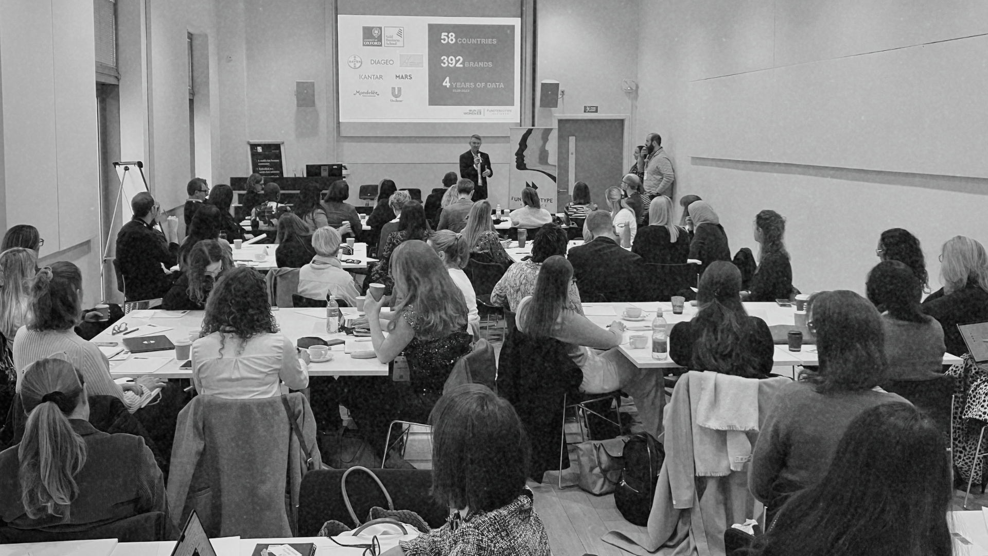 black and white image of people in room watching a person speaking on stage